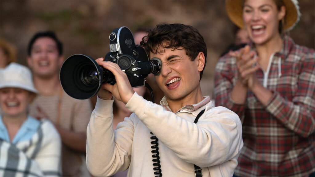 Jovem filma com uma câmera de cinema enquanto outras pessoas observam e comemoram ao fundo, em cena de Os Fabelmans.