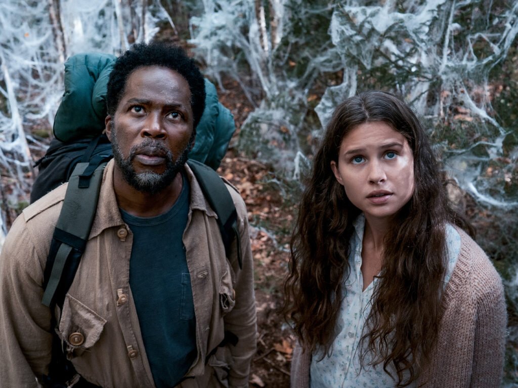Homem e mulher com mochilas observam algo acima deles em uma floresta coberta por teias, em cena de From.