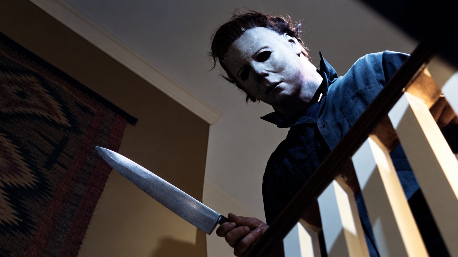Michael Myers usando máscara branca e roupa escura, segurando uma faca no alto de uma escada em uma cena inspirada em Halloween.