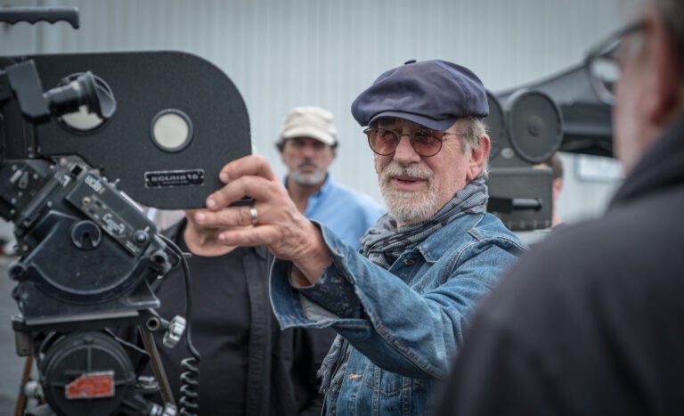 Steven Spielberg opera uma câmera de cinema durante uma filmagem, usando boné, óculos, cachecol e jaqueta jeans.