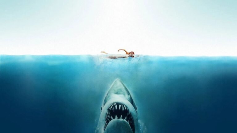 Nadadora na superfície do mar enquanto um grande tubarão se aproxima por baixo da água, em imagem de Tubarão.